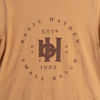 Close-up of the front of the orange shirt. In the center, printed in black, there is the Basil Hayden symbol, with “Basil Hayden” written in a circular fashion around the symbol. The bottom-half of the circle is written “Small Batch”.